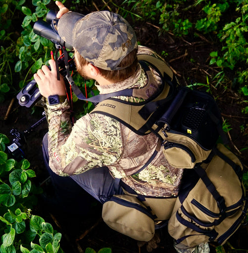 A Man In Camouflage With A Camera In His Backpack And A Binocular In His Hand.