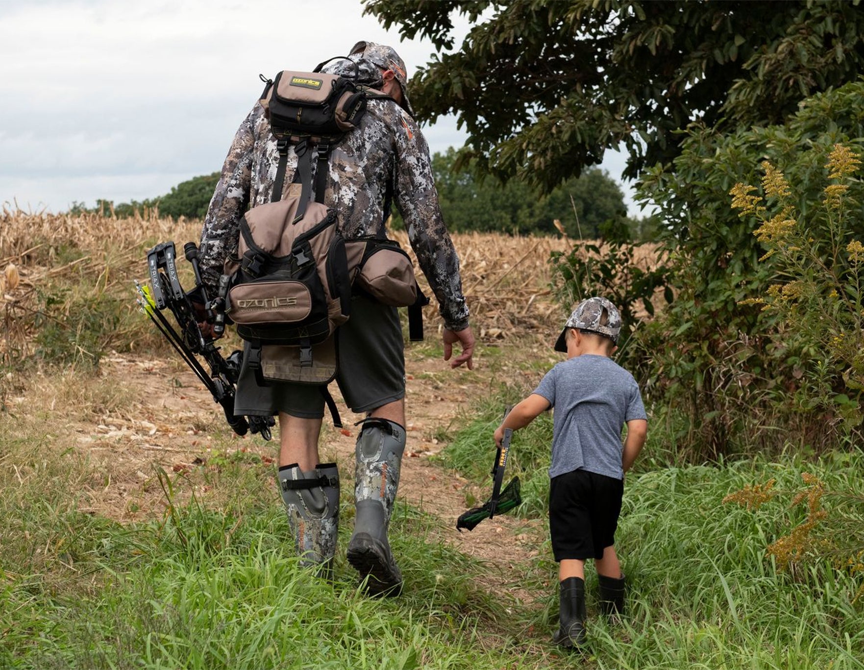 A Man With A Backpack And A Young Boy Walking Through A Field.